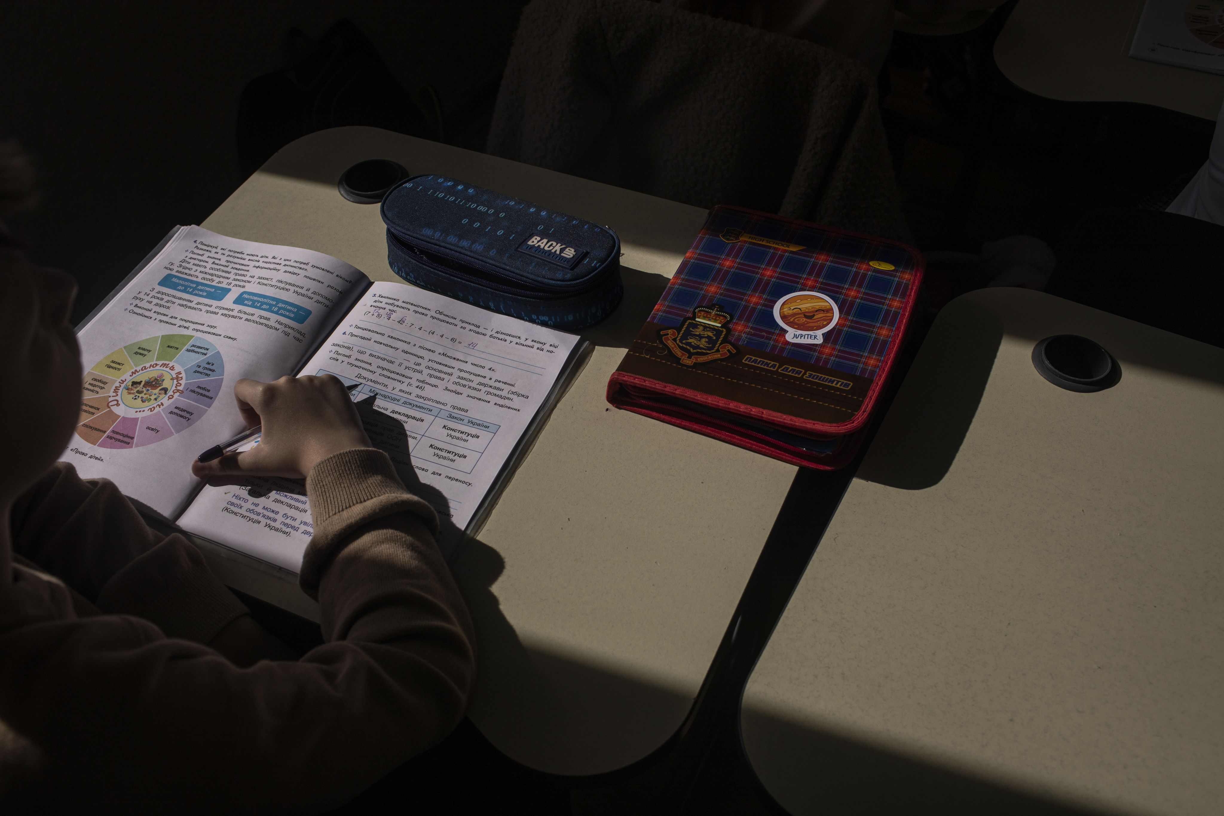 A child holds a pen while reading a textbook in a classroom.